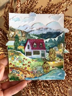 someone is holding up a small card with a house on it and mountains in the background