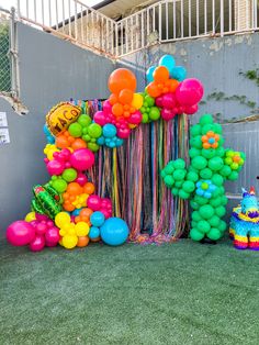 balloons and streamers are arranged on the grass near a fence with a birthday cake