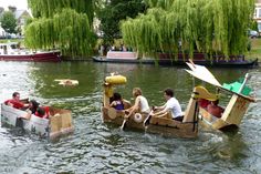 several people are riding in a boat made out of cardboard boxes on the water with trees and boats behind them