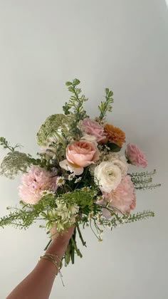 a hand holding a bouquet of flowers on top of a white wall with greenery