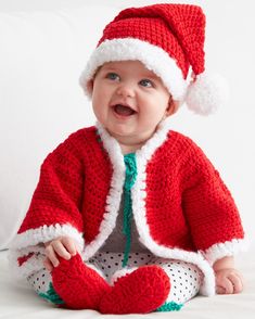 a baby wearing a red and white knitted christmas outfit sitting on a bed smiling at the camera