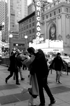 a man and woman kissing on the street in front of a building with people walking by