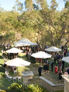 a group of people standing around tables and umbrellas in the grass near some trees