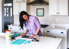 a woman standing at a kitchen counter working on a project with scissors and paint rollers