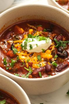 two bowls of chili with sour cream and green garnish on top, sitting on a table