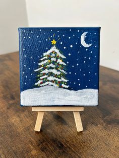 a small christmas tree painted on an easel with the moon and stars in the background