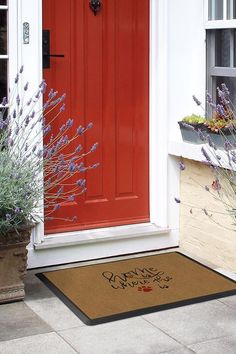 a red front door with a welcome mat on the ground next to flowers and plants