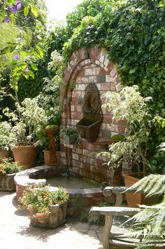 an outdoor fountain surrounded by potted plants and greenery on the side of a brick wall