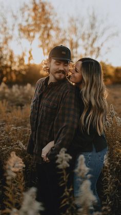 a man and woman standing in tall grass with their arms around each other as the sun sets behind them