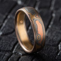 "* Price may vary depending on ring size, please inquire with us directly with your needed size for accurate pricing! This unique Mokume Gane band is shown in the Woodgrain pattern and the Firestorm metal combination, with a low dome profile, and an etched and oxidized finish. Firestorm features 14k red gold, 14k yellow gold, Palladium, and sterling silver.   Pattern: Woodgrain Palette: Firestorm  Finish: Etched and Oxidized Profile: Low Dome Width Shown: 6mm Size Shown: 9.25 Price does NOT include stones or setting fees. We care about customer service and would like to hear from you! Please contact us to help create your treasured item, we take your concerns and requests to heart and will work together to create your unique designs SPECIAL PRICING FOR SETS. CONTACT US WITH YOUR DESIRED WI Hand Forged Adjustable Heirloom Ring, Adjustable Hand Forged Heirloom Ring, Adjustable Hand Forged Fusion Rings, Unique Gold Rings With Patina, Artisan Engraved Rings For Promise, Round Patina Rings For Anniversary, Handmade Brown Ring For Wedding, Etched Round Promise Ring, Adjustable Engraved Round Bands