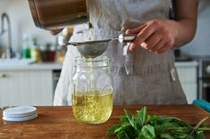 a person pours liquid into a glass jar on a cutting board with herbs in the background