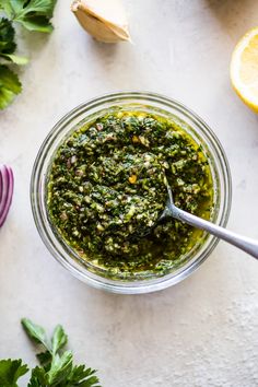 a glass bowl filled with pesto next to sliced lemons and parsley on a white surface