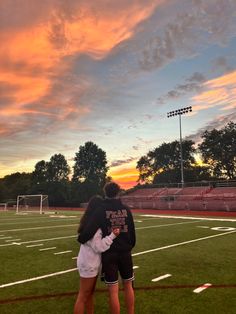 two people standing on top of a soccer field under a cloudy sky at sunset with the sun setting behind them