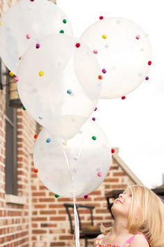 a woman standing in front of a brick building holding white balloons with multicolored confetti on them