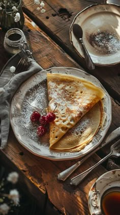 pancakes with berries and powdered sugar sit on a plate next to silverware, cups and saucers