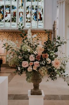 a vase filled with lots of flowers on top of a white pedestal in front of a stained glass window