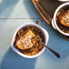 two white bowls filled with beans and toast on top of a blue tablecloth next to a serving tray