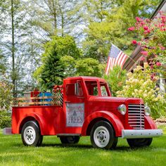 an old red truck is parked on the grass in front of some trees and flowers