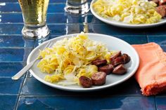 two white plates topped with pasta and sausage next to glasses of beer on a blue tiled table