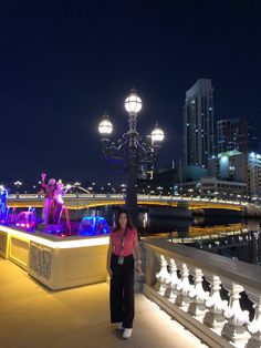 a woman standing in front of a fountain with lights on it and buildings in the background