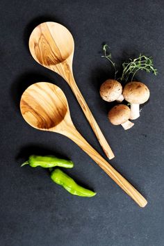 three wooden spoons and two green peppers on a black surface with mushrooms in the background