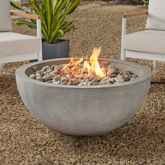 a fire pit sitting on top of a gravel ground next to two chairs and a potted plant