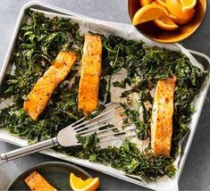 two pans filled with fish and greens next to orange wedges