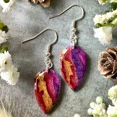 two pairs of earrings with purple and red designs on them sitting next to white flowers