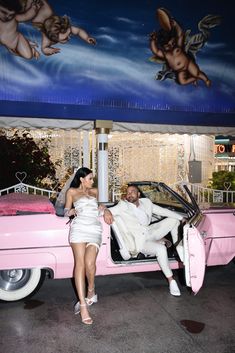 a man and woman sitting on the back of a pink car in front of a mural