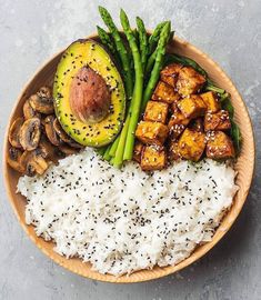 a wooden bowl filled with rice, asparagus and tofu next to sliced avocado