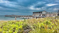 the beach is next to a pier and some grass in front of it on a cloudy day