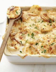 a casserole dish with potatoes and parmesan cheese on top, next to a wooden spoon