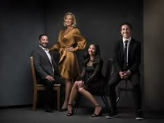 three people are posing for a photo in front of a dark background, one is wearing a black suit and the other wears a gold dress