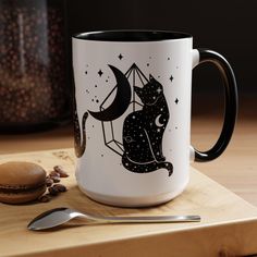 a black cat sitting on top of a white coffee mug next to a spoon and cookie