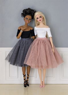 two barbie dolls standing next to each other in front of a gray and white wall