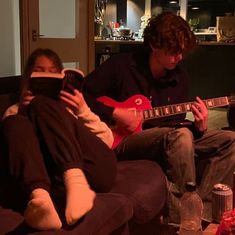 two people sitting on a couch playing guitar and drinking water in the living room while one person is holding an mp3 player