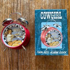 a red alarm clock sitting on top of a wooden table next to a copy of cowgirl