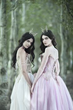 two women dressed in gowns standing next to each other with trees in the background