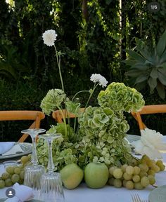 an arrangement of flowers, fruit and wine glasses on a dining table with greenery in the background