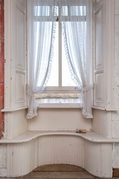 an old window with white curtains and a bench in front of the window sill