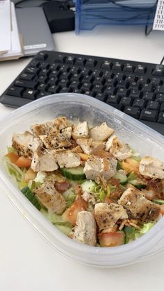 a salad in a plastic container sitting next to a computer keyboard on a white desk