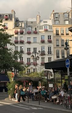 many people are sitting at tables in front of tall buildings with flower boxes on the windows