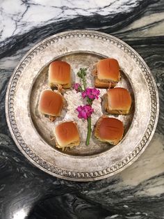 small sandwiches are arranged on a silver platter with pink flowers in the middle and purple petals sticking out of them