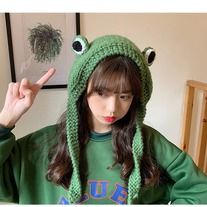 a woman wearing a green frog hat with two eyes on it's face, making the peace sign