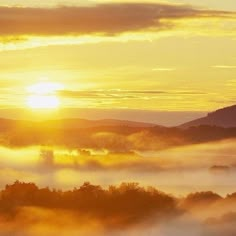 the sun is setting over some hills and trees in the foggy valley with low lying clouds