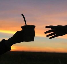 two hands reaching out towards each other with a cup in front of the sun setting