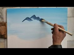 a person holding a paintbrush in front of a painting with mountains on the side