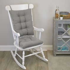 a white rocking chair with grey cushions next to a gray side table and glass cabinet