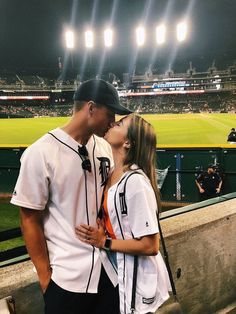 a man and woman kissing in front of a baseball field at night with bright lights