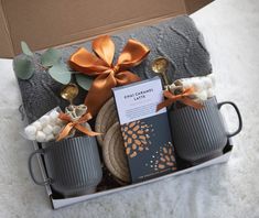 two coffee mugs with spoons in a gift box on a white blanket next to a tea towel